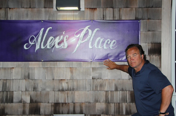 A man standing next to a sign that says " alex 's place ".